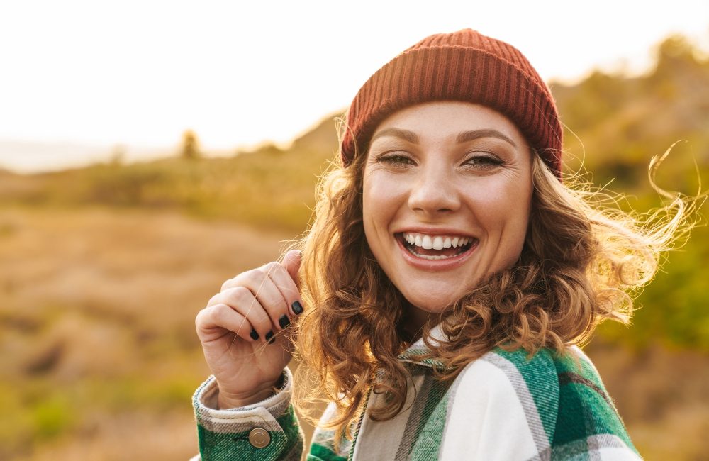 Image of joyful young caucasian woman wearing hat and plaid shirt smiling while walking outdoors